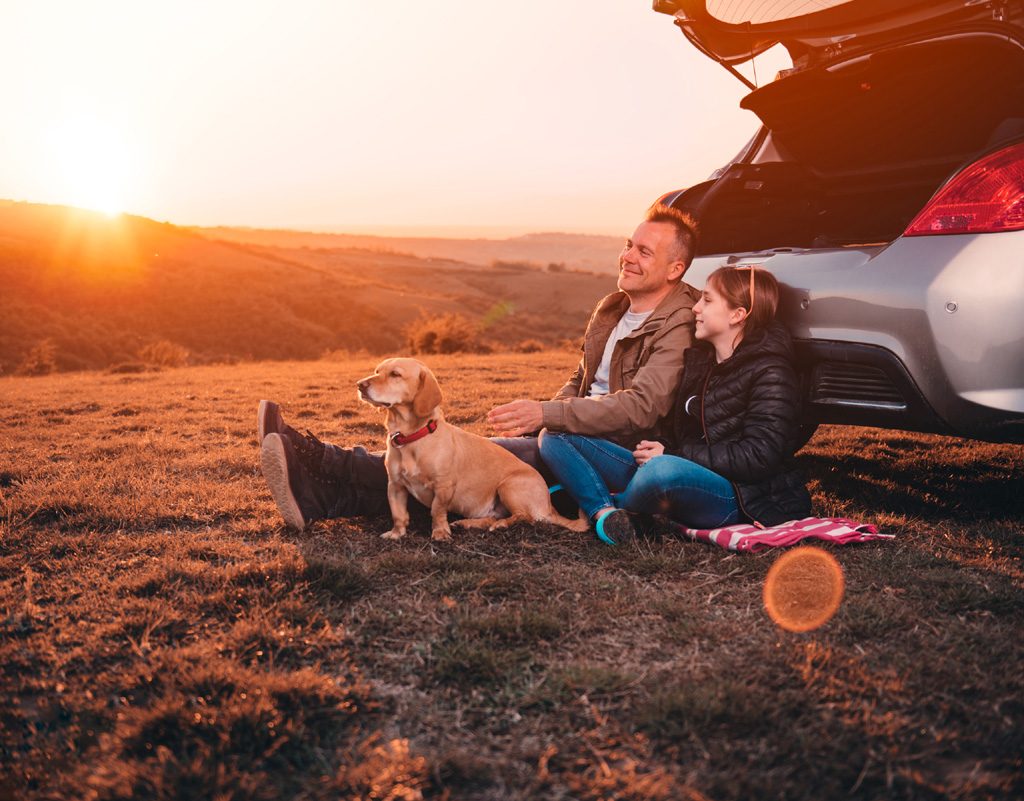 man, girl and dog sitting beside a car