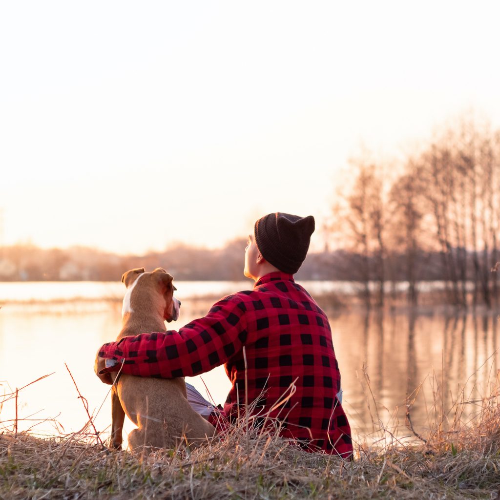 A man sits with his arm around his dog in front of a lake with their backs to the camera