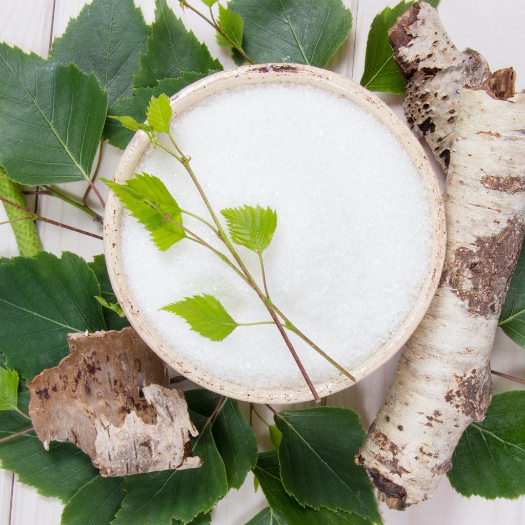 a bowl of white sugar-like xylitol sits in the middle of a spread of green leaves, next to pieces of birch wood