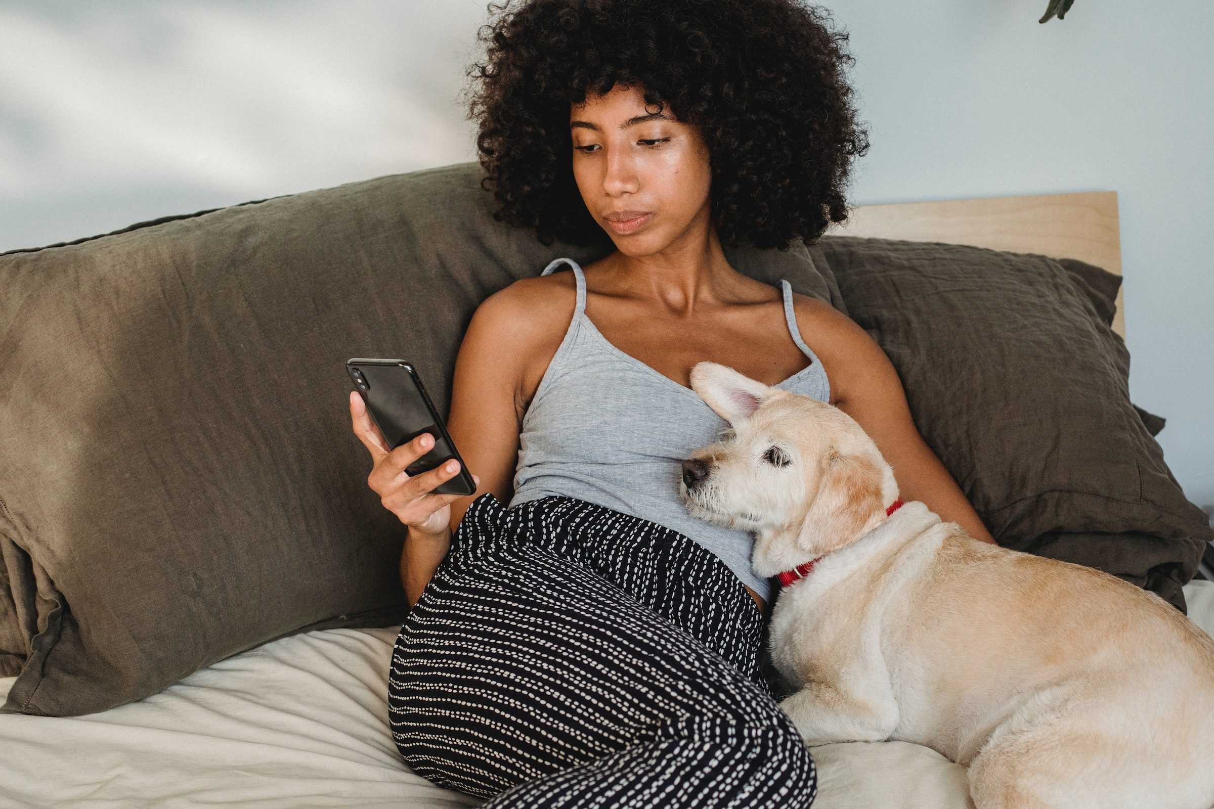 a woman reclines in bed with her dog leaning into her side while she looks at her smartphone