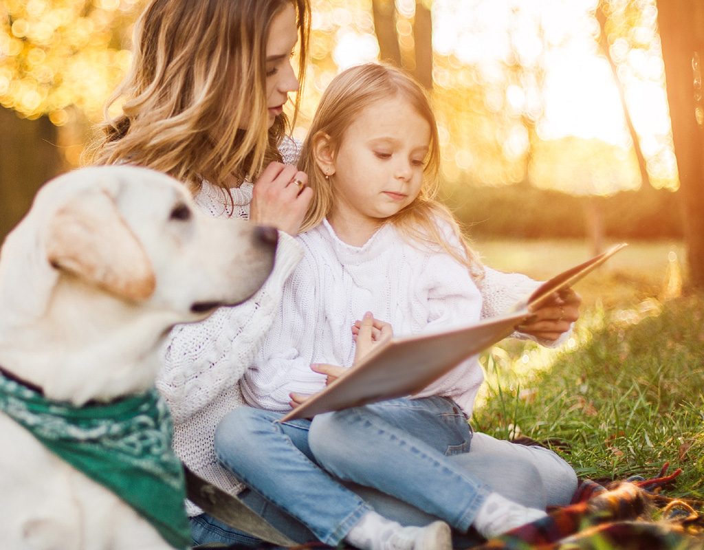 Woman reading book with child while dog sits besides them.