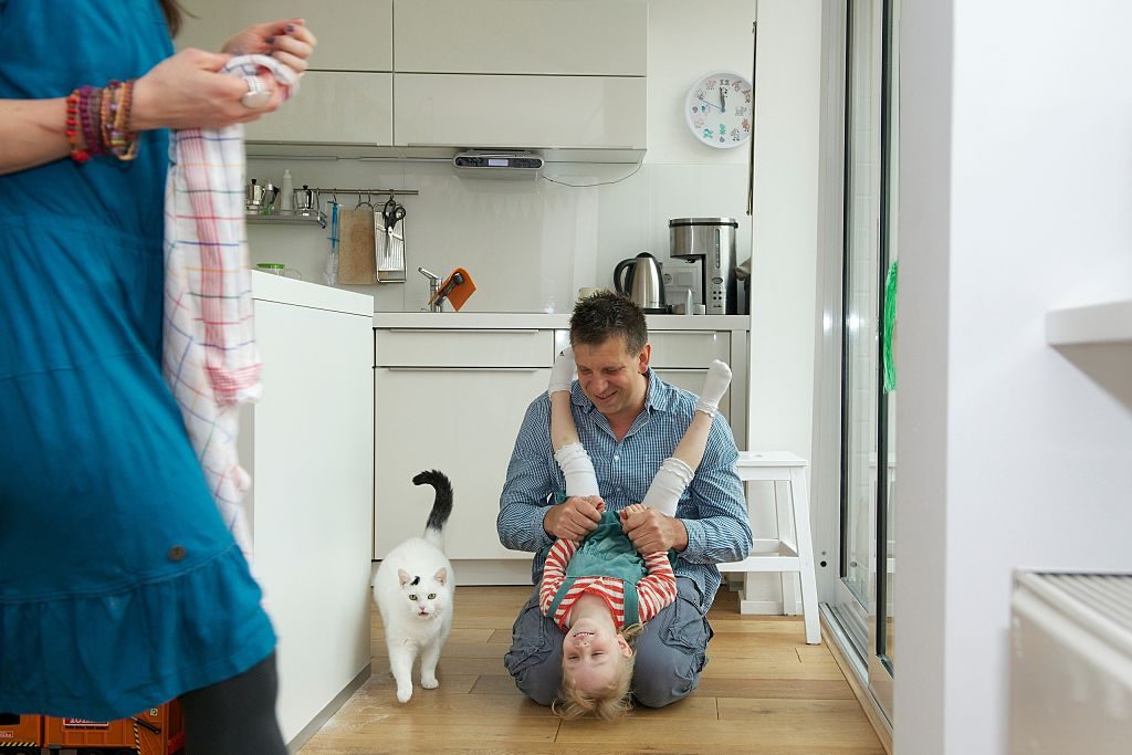 A father plays with a young child in a kitchen with a cat and the mother look on.