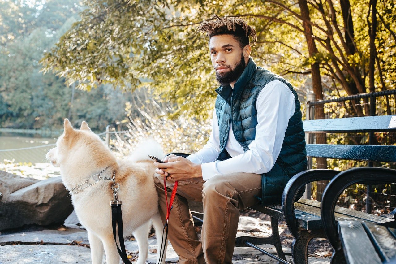 A man sits on a park bench using his phone while his leashed dog stands beside him.