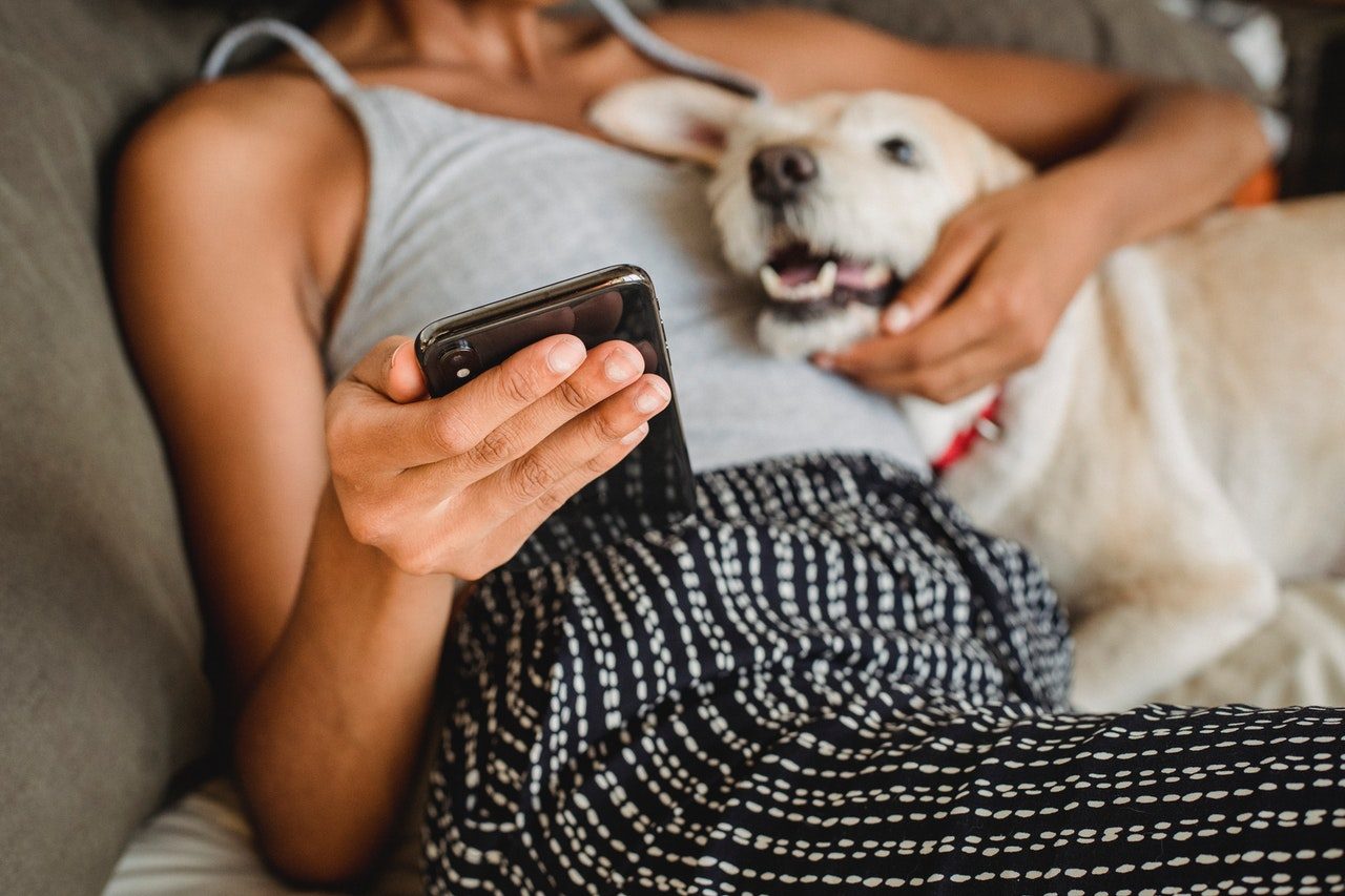 A woman in a gray tank top uses her phone while cuddling a beige dog.