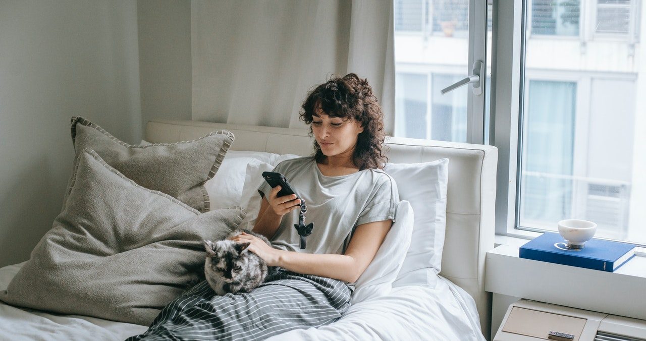 A woman lounges in bed using her phone while a calico cat stretches out on her lap.