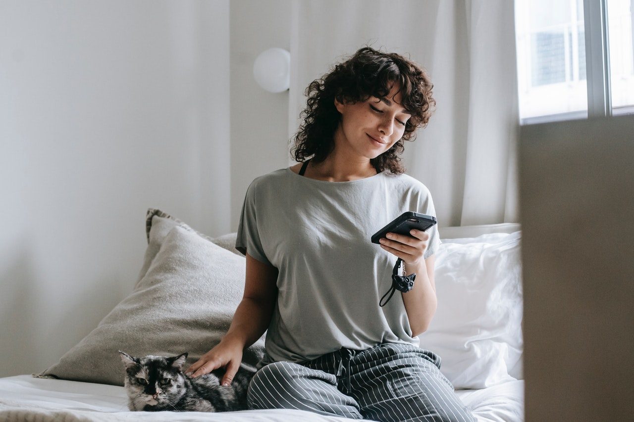 A woman in a gray t-shirt pets a calico cat while she uses her phone.