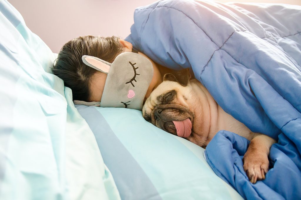 A woman wearing a sleep mask over her eyes snuggles a Pug with his tongue out in bed