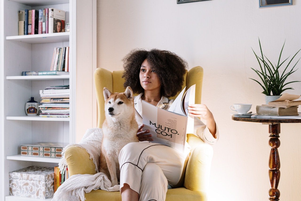 A Shiba Inu sits in a yellow chair with a woman reading a newspaper.