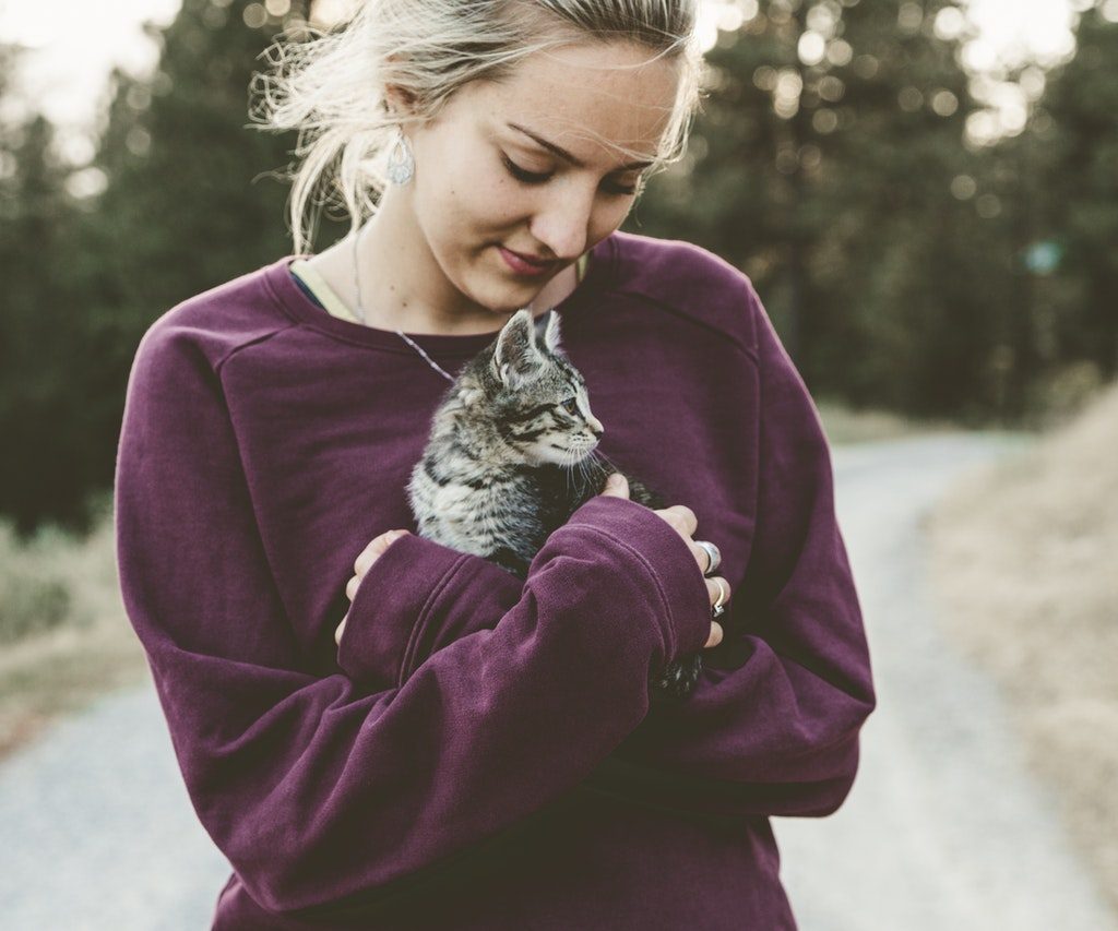 A blonde woman wearing a purple sweatshirt holds a tabby kitten in her arms.