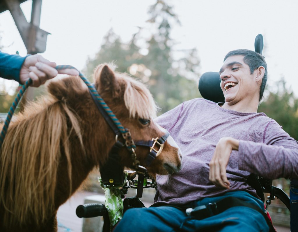 Man in a wheelchair laughing as he pets a horse.