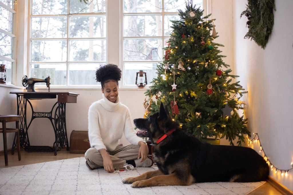 A woman wearing a cream turtleneck sweater sits beneath a Christmas tree with her German shepherd.