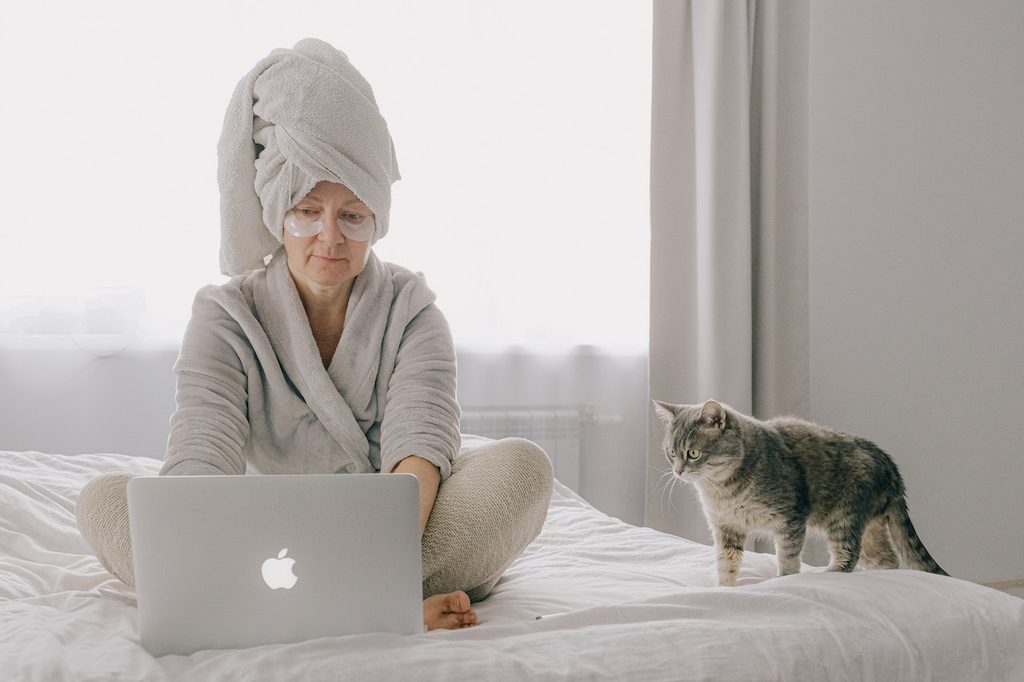 A woman wearing eye patches and a pale gray robe works on her laptop on a bed with a white comforter. She has her hair wrapped in a towel, and a gray cat stands beside her.