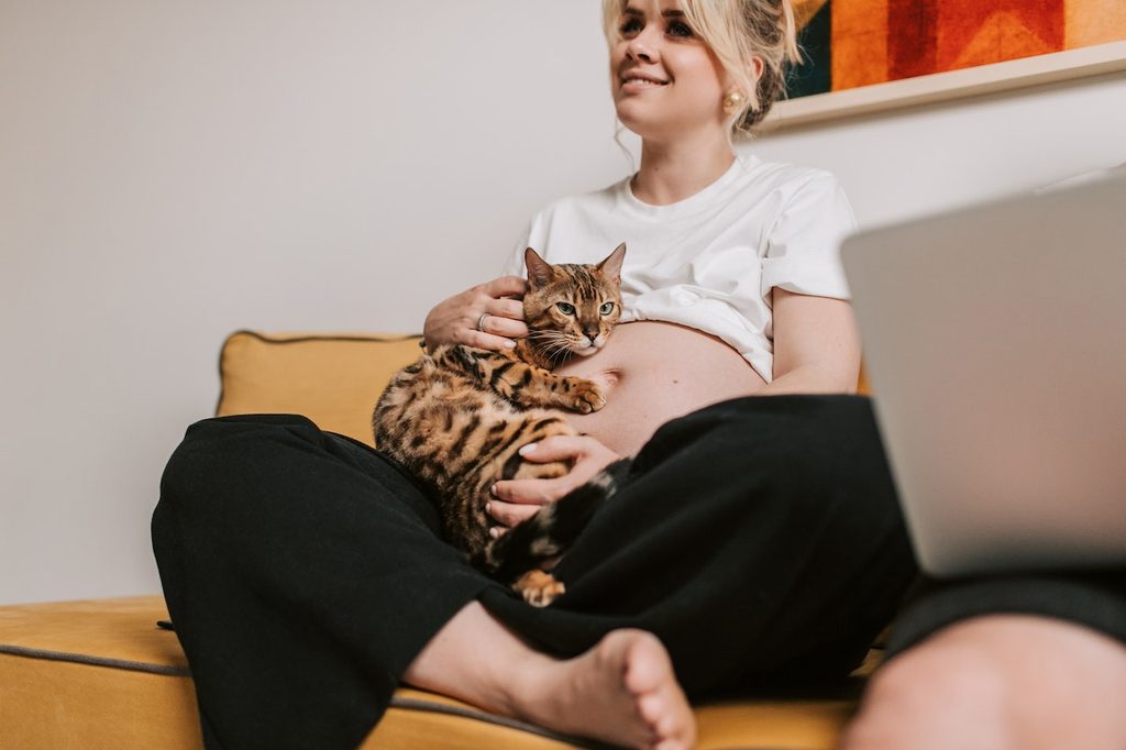 A pregnant woman sits cross-legged on a yellow sofa as she holds a Bengal cat