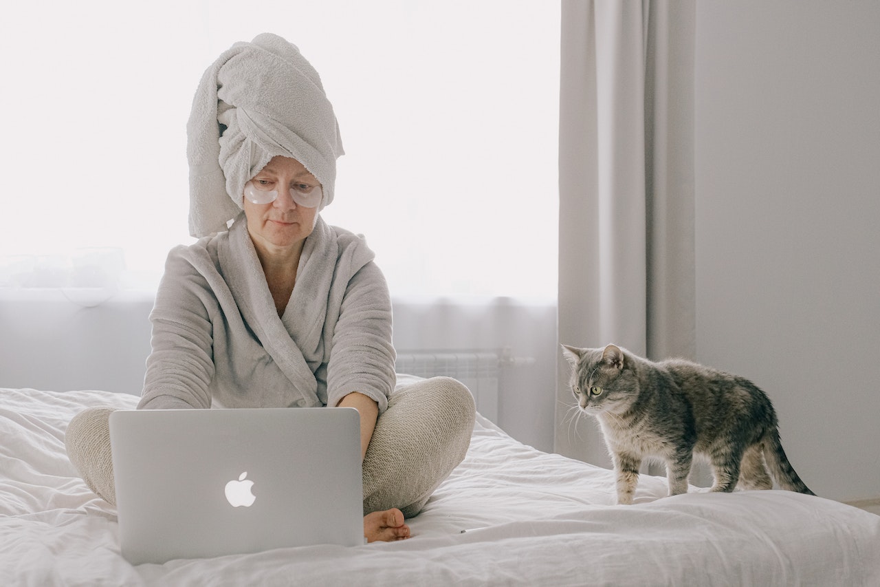 A woman wearing eye patches and a pale gray robe works on her laptop on a bed with a white comforter. She has her hair wrapped in a towel, and a gray cat stands beside her.