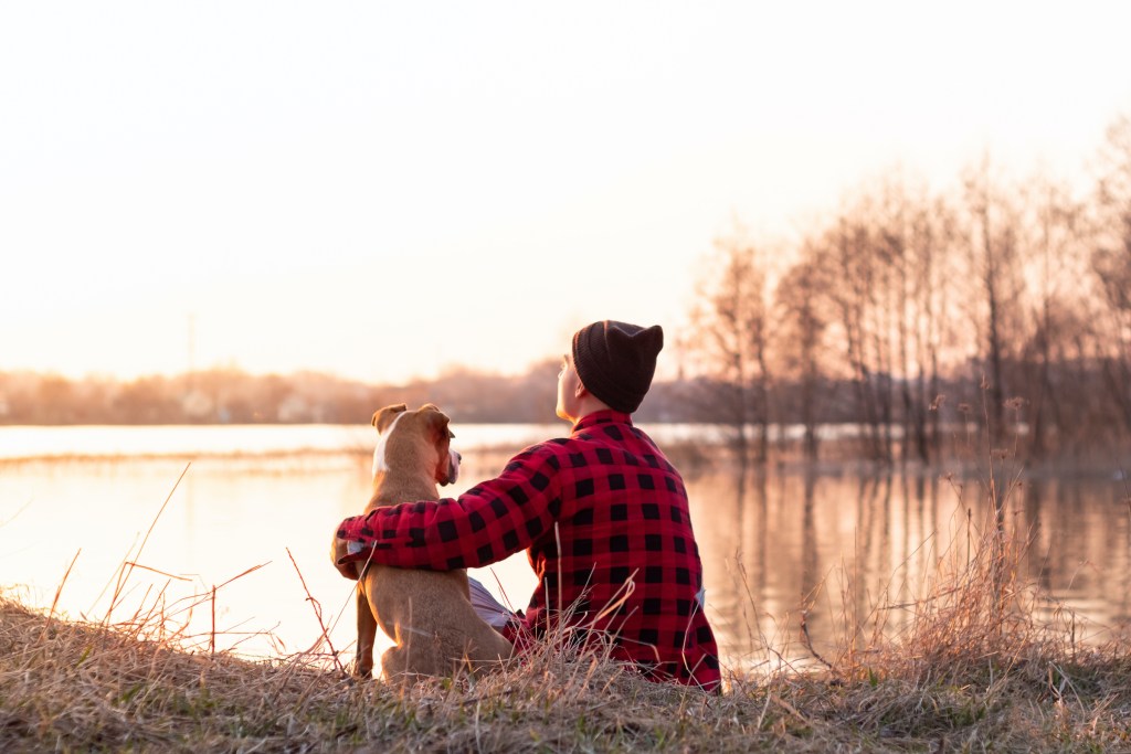 A man sits with his arm around his dog in front of a lake with their backs to the camera