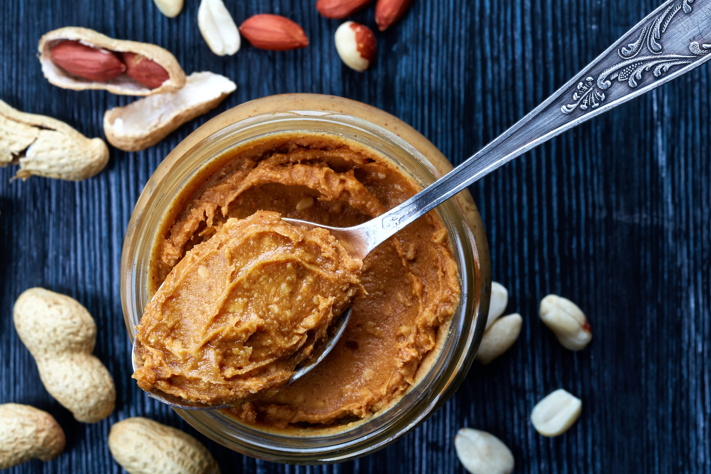 a spoonful of peanut butter sits on top of a peanut butter jar with cracked peanuts sprinkled on the table around it