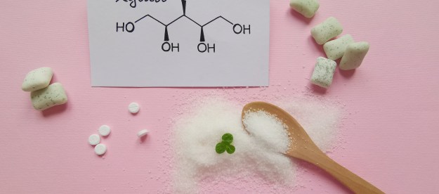on a pink background: a wooden spoon in a pile of Xylitol powder, a notecard with the chemical structure of xylitol, pieces of chewing gum, and small circular leaves