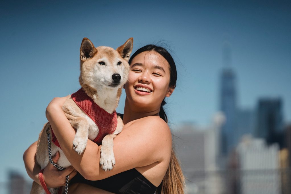 A smiling young woman holds a shiba inu in her arms in front of a city in the background