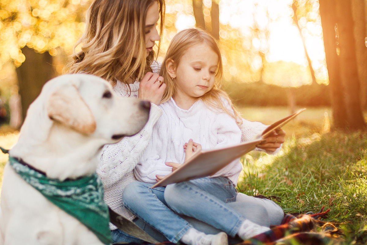 Woman reading book with child while dog sits besides them.