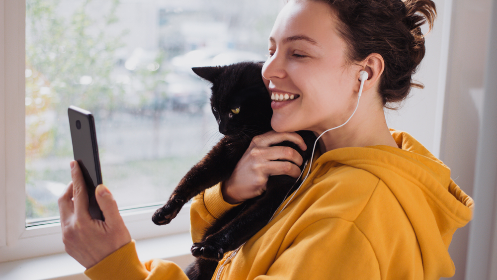 A woman in a golden yellow hoodie holds a black cat while using her smartphone.