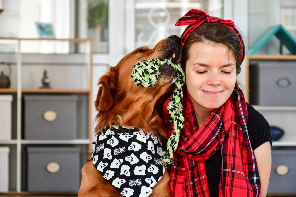 A Golden Retriever wearing a ghost bandana snuggles a brown-haired woman wearing a red scarf.