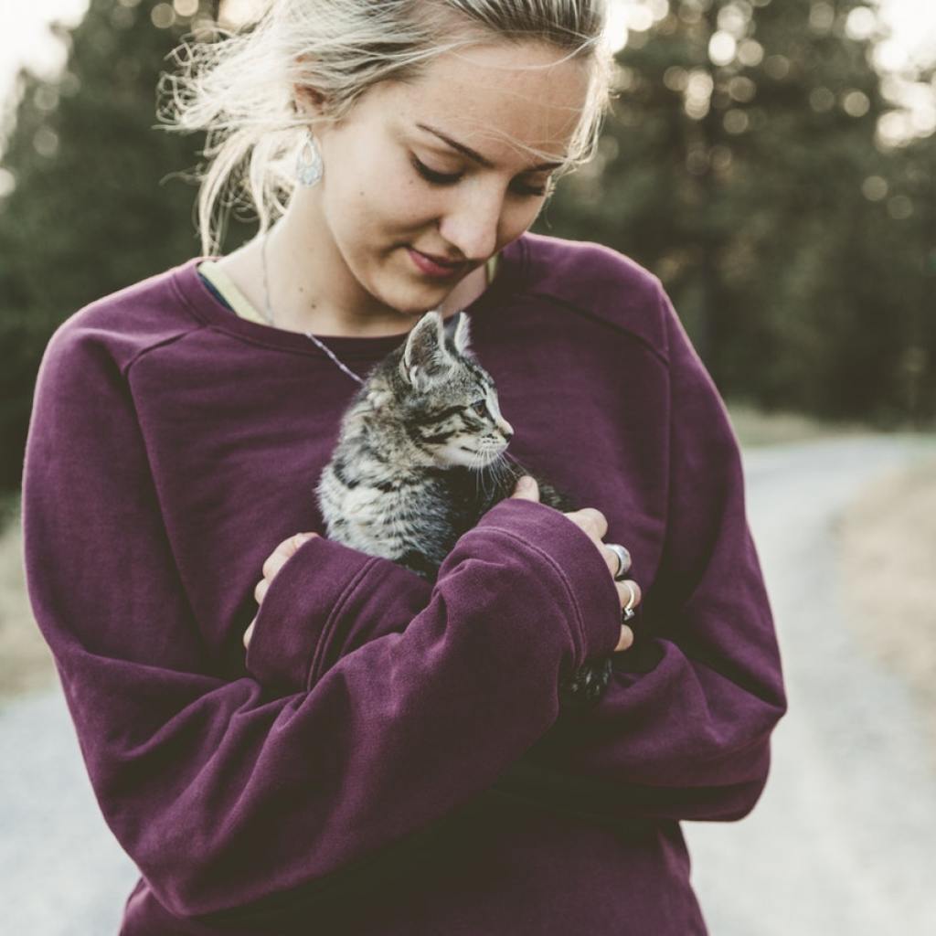 A blonde woman wearing a purple sweatshirt holds a tabby kitten in her arms.