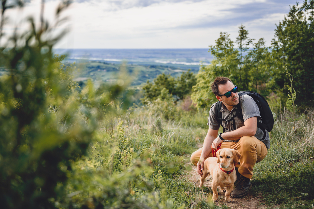 A man wearing sunglasses crouches on a hiking trail with a small brown dog.