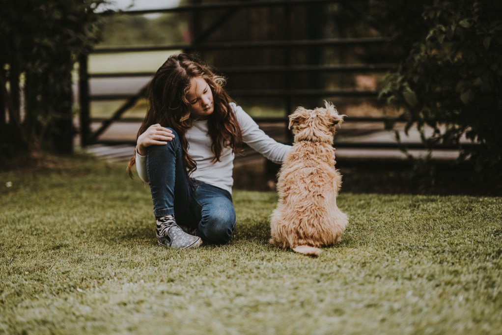 A girl sits outside on the grass and pets the dog sitting next to her