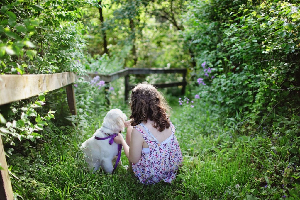 A little girl and a puppy sit on the grass in a lush green outdoor area