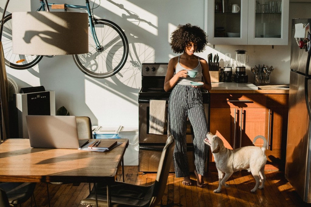 a dog in the kitchen looking up at woman drinking coffee